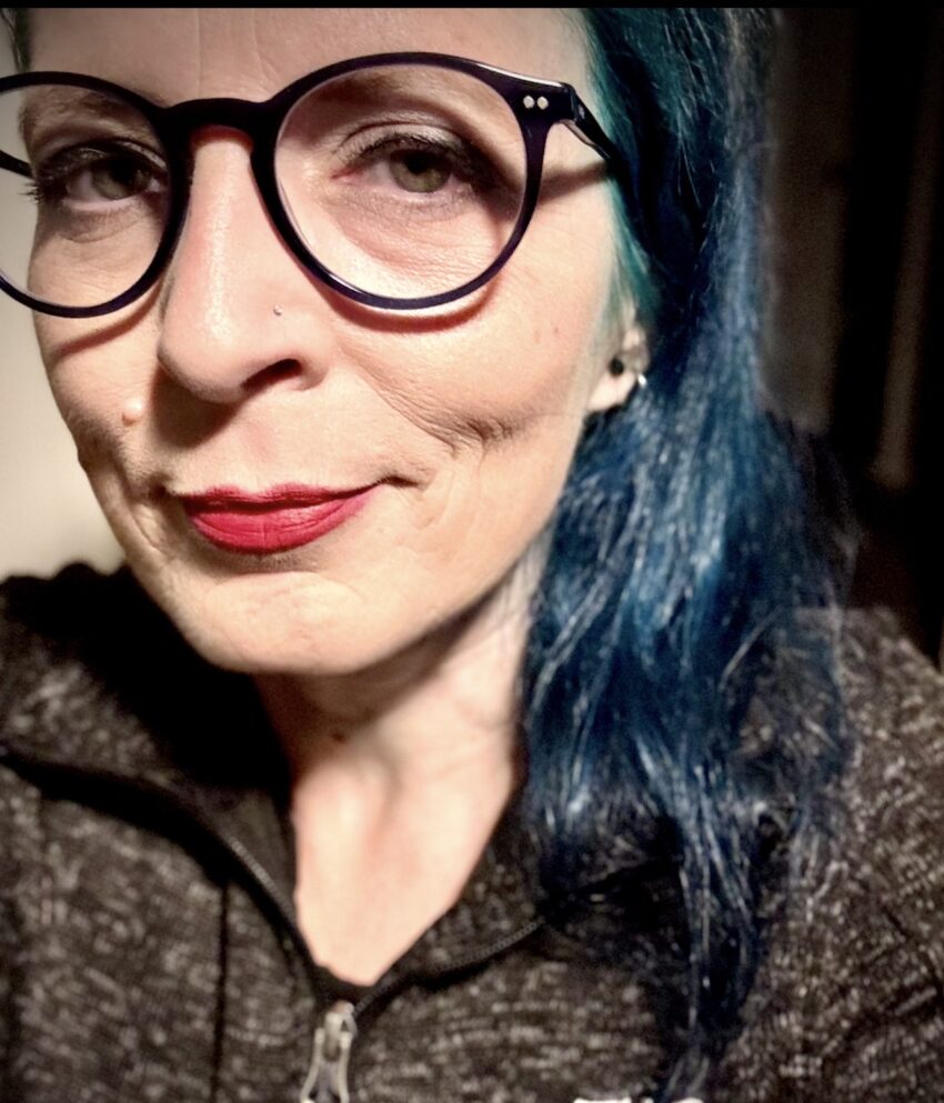 A woman with blue hair, wearing glasses and a grey jacket is smiling at you